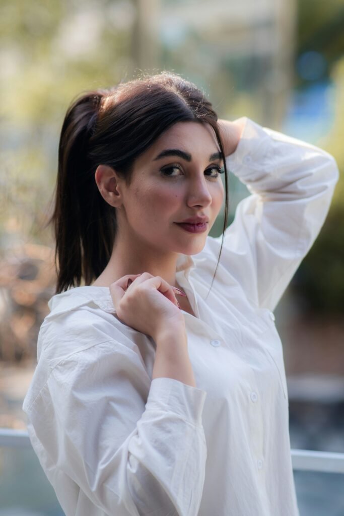 mulher com cabelos lisos longos pretos olhos escuros uma das mãos segurando os cabelos camisa manga comprida branca