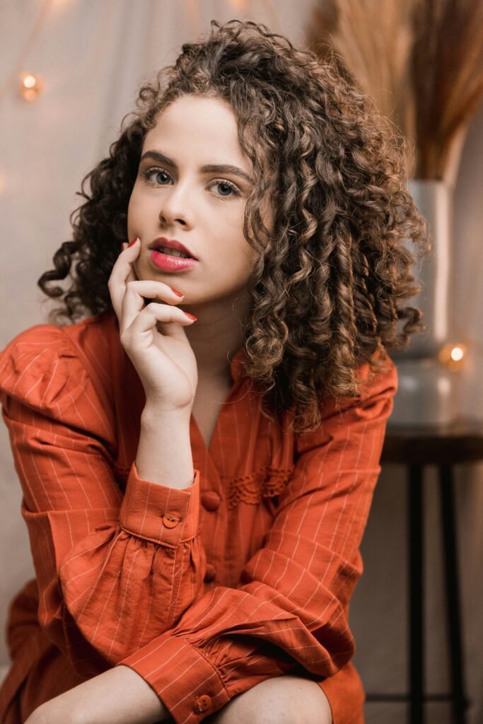 mulher maquiada cabelos longos cacheados cor castanho blusinha estilosa cor laranja uma das mãos no queixo