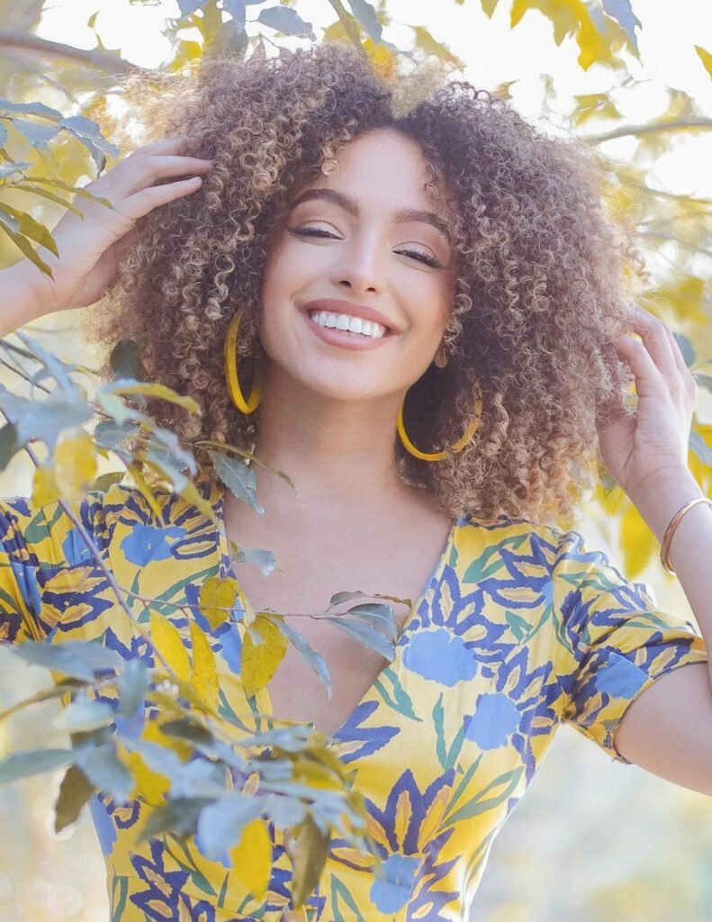 foto de mulher morena linda sorrindo cabelos encaracolados grandes brincos argolas amarelos, blusinha amarela florida duas mãos nos cabelos