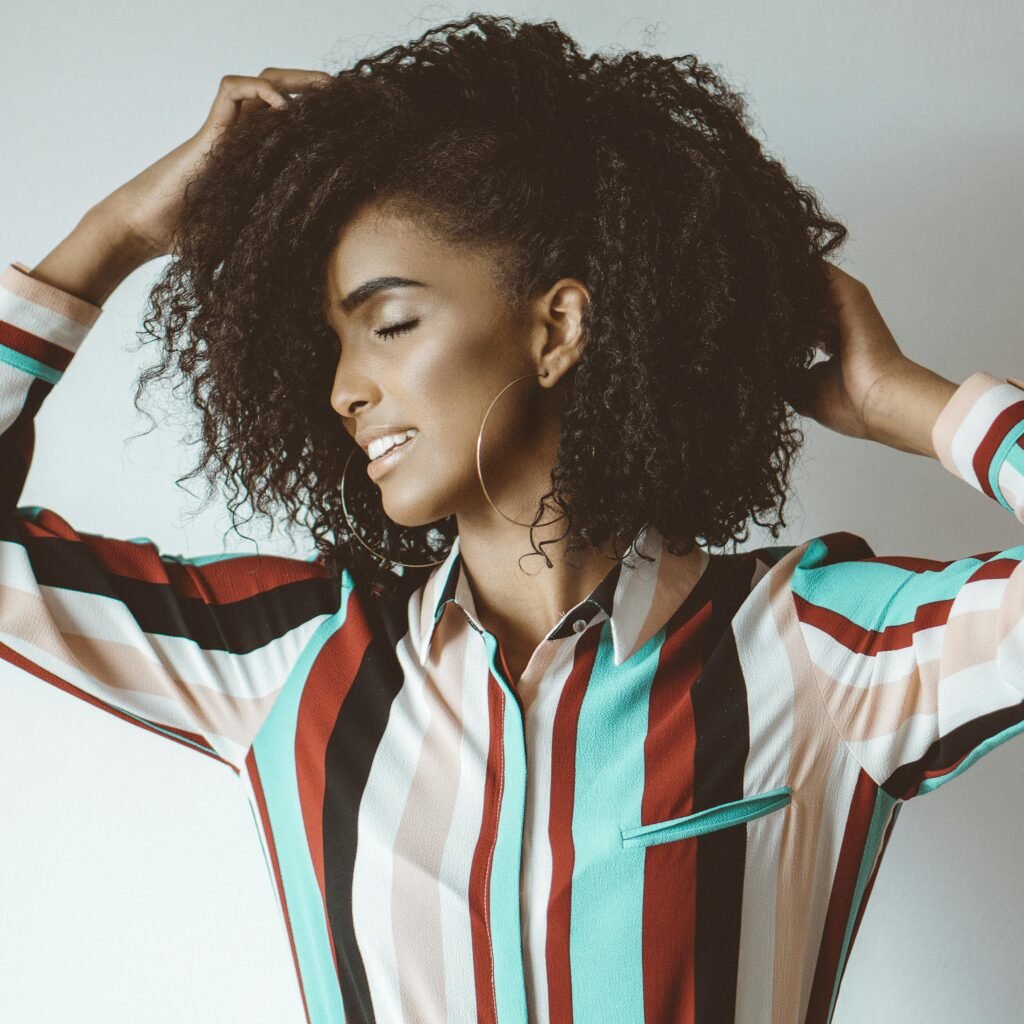 foto de mulher morena linda leve sorriso brincos argolas grandes camisa social listrada duas mãos nos cabelos