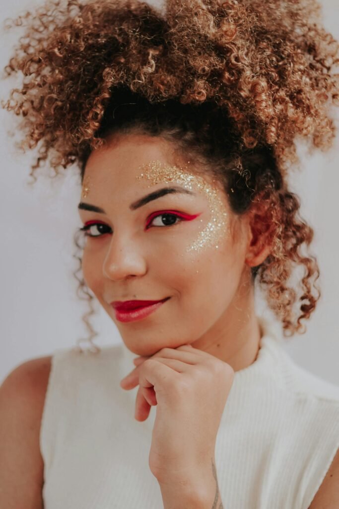 mulher maquiada leve sorriso uma das mãos no queixo cabelos cacheados volumosos preso em penteado coque grande blusinha branca