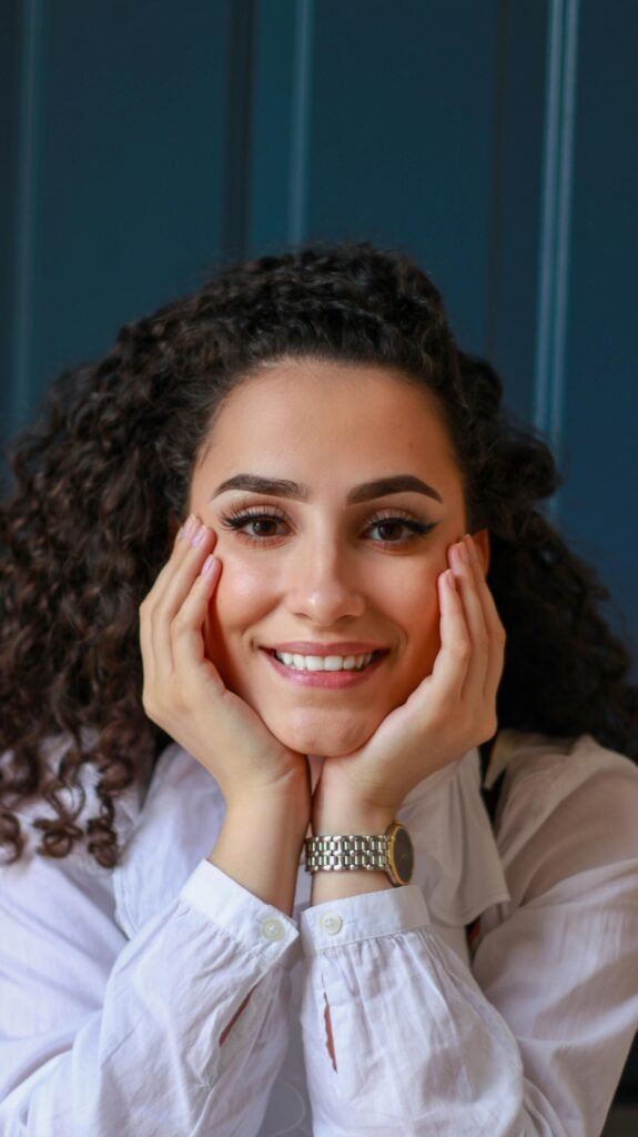 foto de perfil de mulher branca sorrindo cabelos longos pretos cacheados esta com as duas mãos segurando o rosto