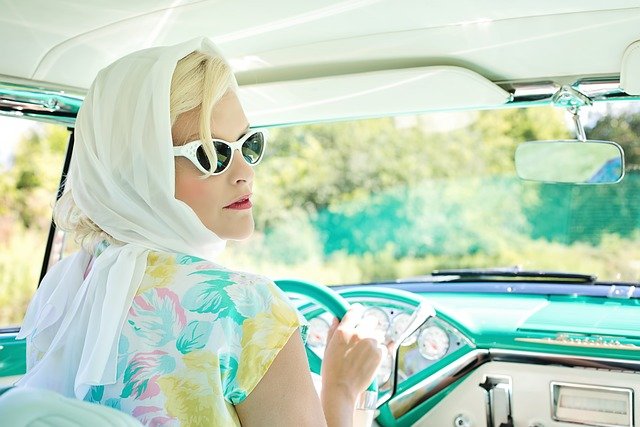 foto retro de uma mulher loira com oculos dirigindo um carro antigo na cor azul