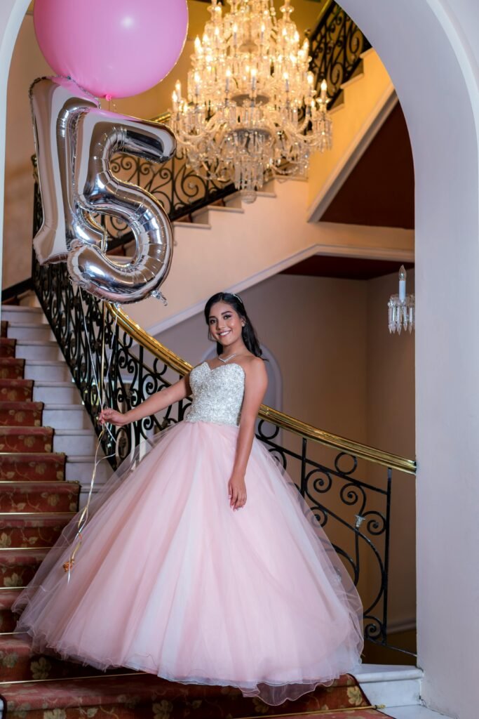 menina sorrindo comemorando seus 15 anos vestido longo na cor rosa claro, segurando dois baloes em forma de numero 15, esta em uma escadaria tipo imperial