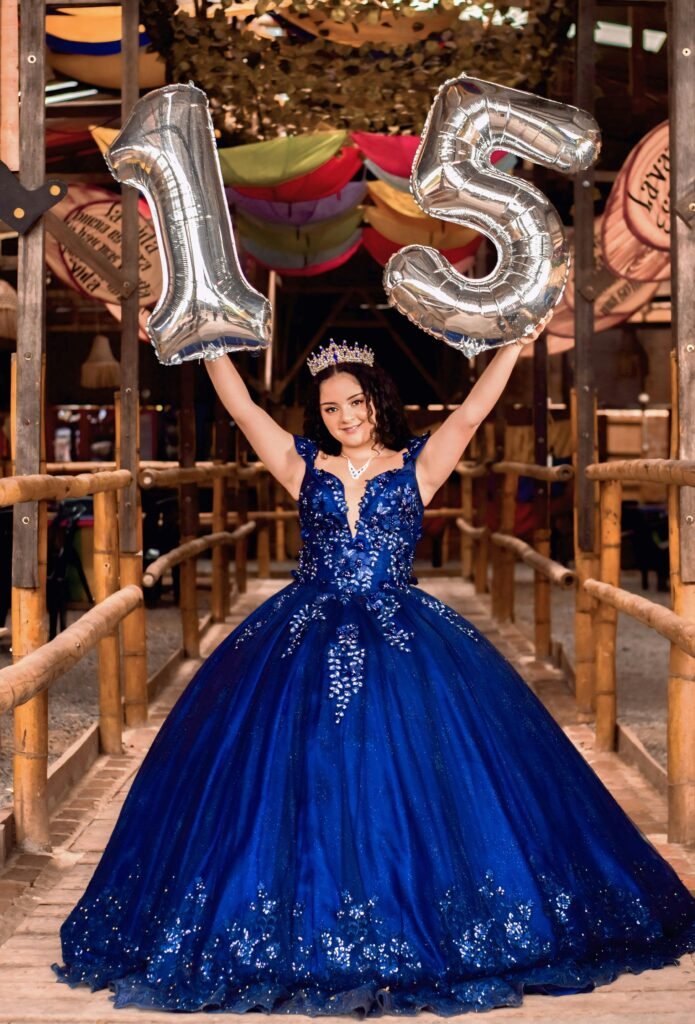 menina sorrindo comemorando seus 15 anos segurando dois baloes em forma de números 15 vestido de princesa na cor azul escuro leve sorriso e coroa de princesa