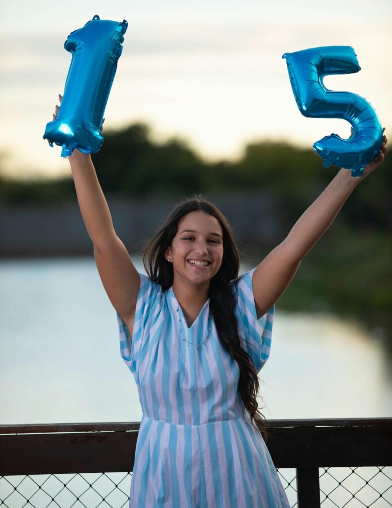 menina sorrindo comemorando seus 15 anos, bracos levantados ao alto segurando dois baloes em forma de numero 15, vestido listrado branco e azul