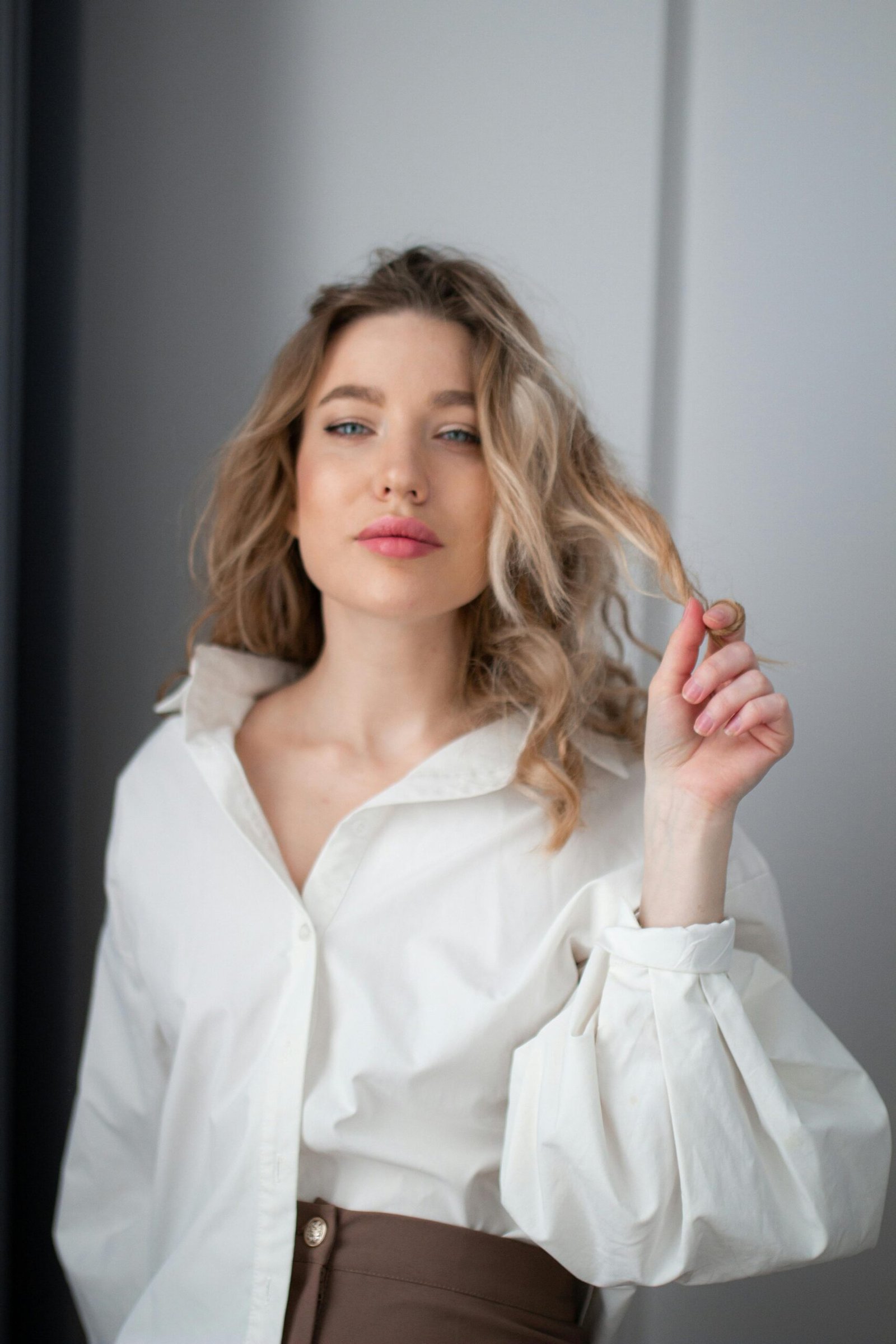 Foto de mulher loira cabelos longos ondulados soltos leve maquiagem blusinha social branca uma das mãos nos cabelos