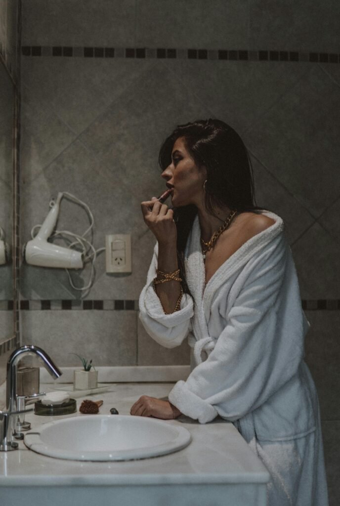 foto de mulher morena cabelos lisos compridos no banheiro com roupão branco passando batom