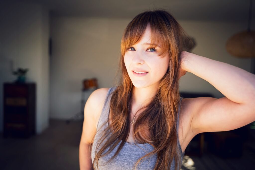 mulher cabelos lisos longos castanhos com franja olhos azuis uma das mãos na cabeça blusinha cinza