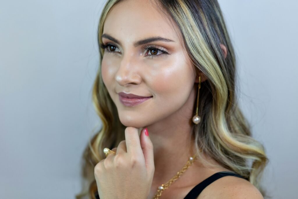 loira maquiada leve sorriso olhos castanhos cabelos lisos longos soltos brincos grandes perola anel perola colar dourado uma das mãos no queixo
