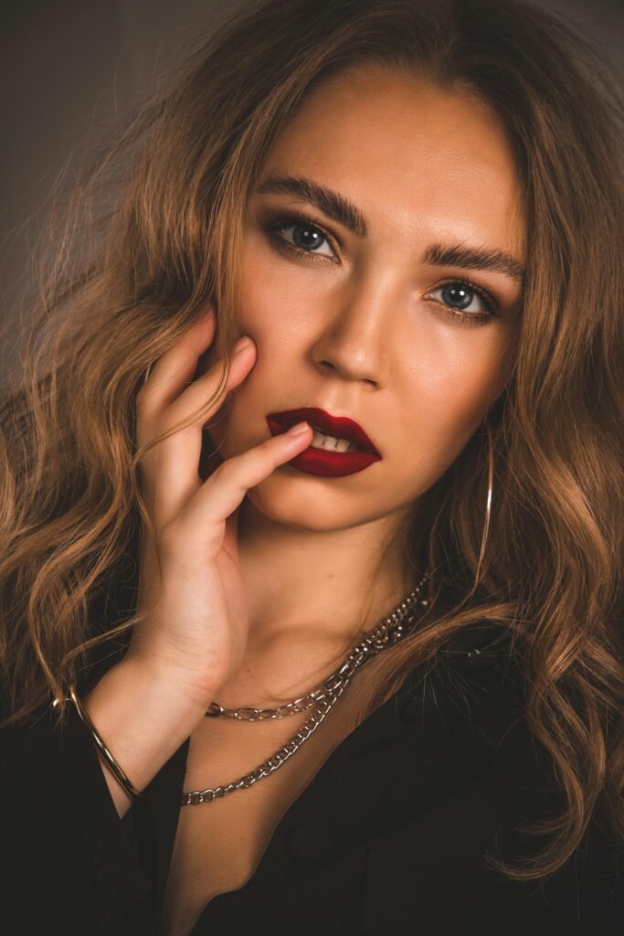 mulher maquiada batom vermelho olhos azuis cabelos lisos longos soltos uma das mãos no rosto brincos argolas grandes colares de prata
