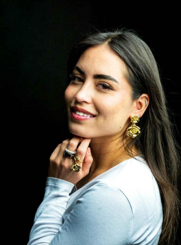 mulher sorrindo olhos castanhos claros batom vermelho cabelos lisos pretos soltos brincos dourados grandes anéis nos dedos uma das mãos no queixo blusinha branca