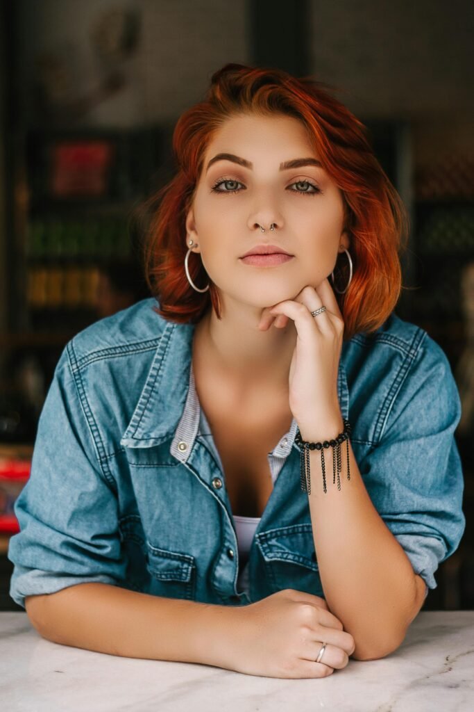 mulher jovem branca seria com percingi no nariz brincos grandes argola olhos verdes claros cabelos médios vermelhos, uma das mãos segurando o queixo tipo pensando