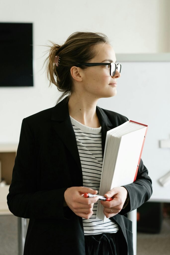 mulher empresaria de oculos branca cabelo preso leve sorriso segurando uma pasta casaco preto segurando uma caneta