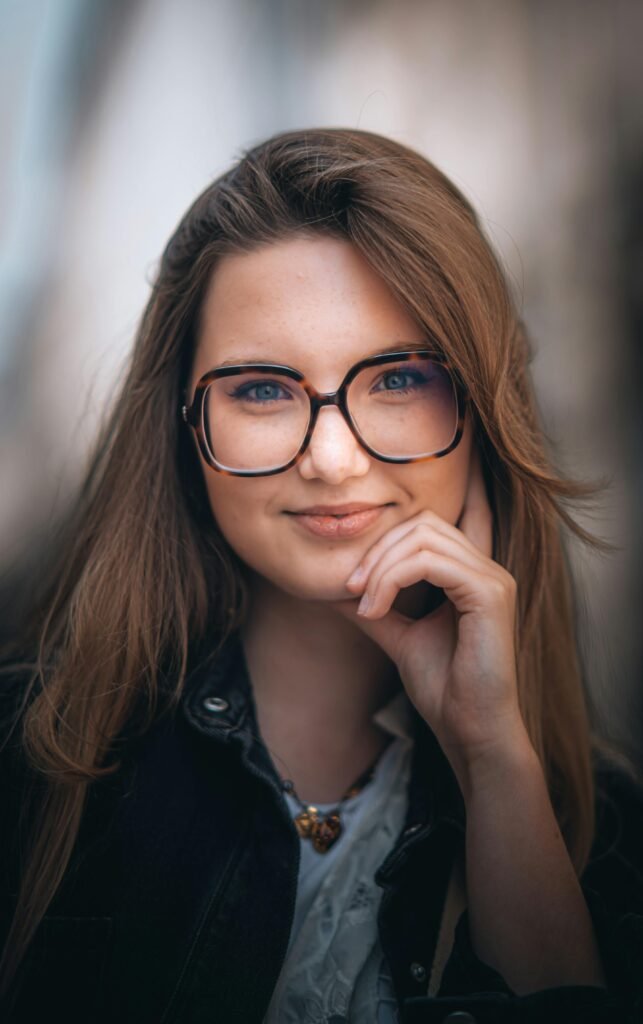 uma mulher branca com a mão no queixo, leve sorriso, de óculos, cabelos longos lisos, jaqueta preta