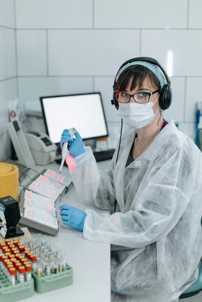 foto de uma mulher de óculos mascara na boca fones com jaleco de proteção fazendo pesquisa em laboratório