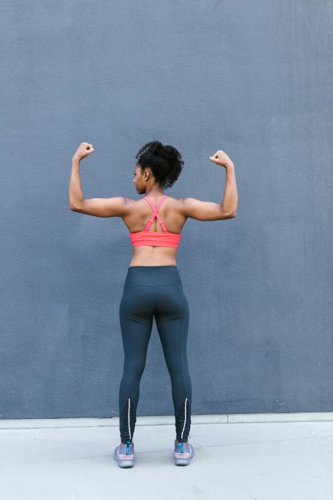 foto de mulher morena com roupa de academia esta de costas cabelos encaracolados, fazendo pose de força com os bracos
