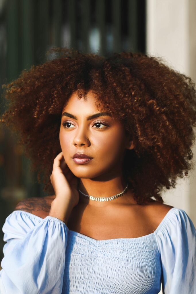 mulher maquiada cabelos crespos grandes olhos castanhos escuros colar de prata batom marrom tatuagem no ombro blusinha estilosa branca