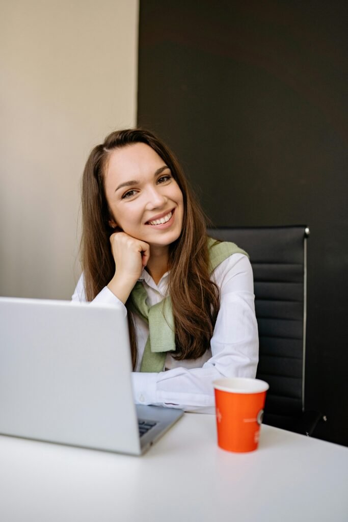 mulher branca sorrindo cabelos longos lisos, com a mão fechada segurando o queixo sentada defronte ao notebook camisa branca