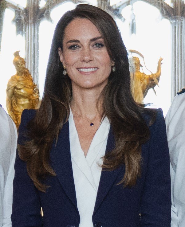 foto da Princesa kate Middleton sorrindo cabelos castanhos lisos longos terno zaul camisa social branca colar pequeno