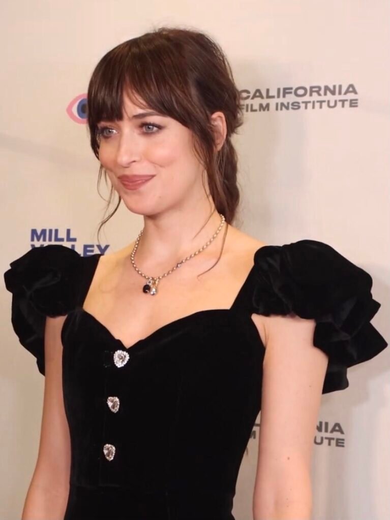 foto de Dakota johnson leve sorriso linda cabelo lisos presos penteado franja colar dourado vestido de festa estiloso preto