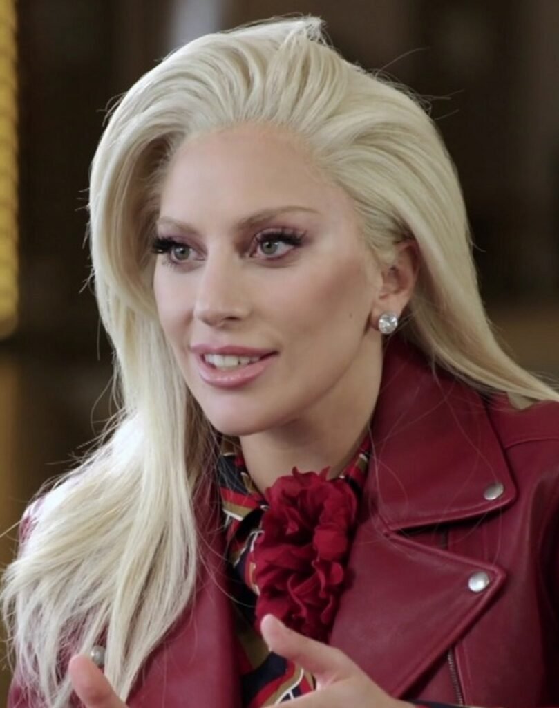 lady gaga sorrindo maquiada linda olhos verdes claros brinco pequeno de diamante cabelos compridos loiros lisos jaqueta de couro vermelha