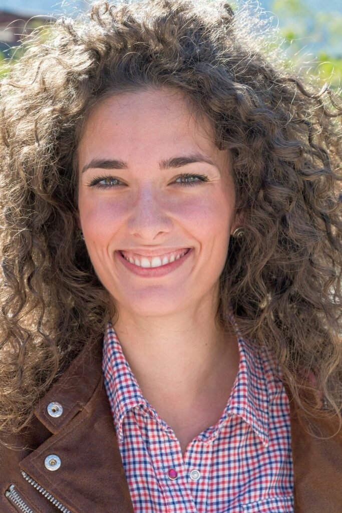 mulher sorrindo olhos castanhos claros cabelso longos volumosos cacheados castanhos claros camisa social xadrez jaqueta de couro marrom