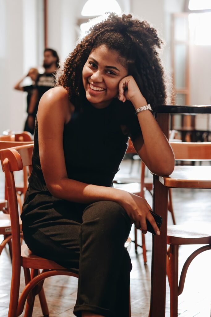 mulher sorrindo cabelos crespos grandes lindos blusinha preta uma das mãos no rosto relógio no braco