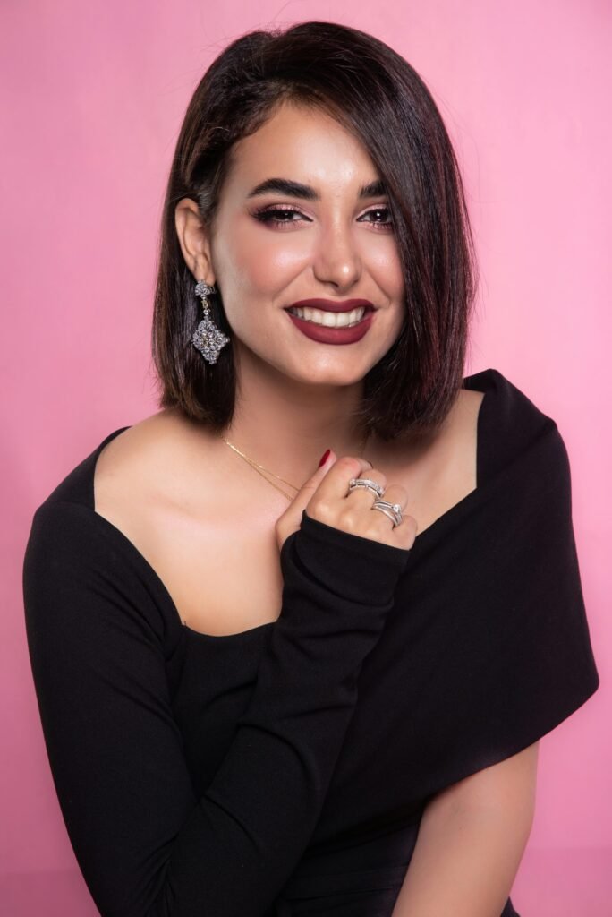 mulher branca linda sorrindo batom roxo maquiada brinco grande triangulo prata vestido preto estiloso unhas vermelhas anéis pratas nos dedos