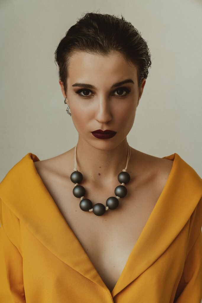 mulher seria maquiada batom roxo dois brincos pequenos argolas em uma orelha colar artesanal com bolas vestido estiloso amarelo
