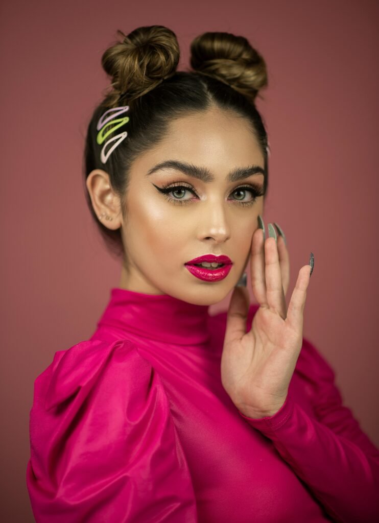 mulher maquiada olhos verdes claros batom rosa cabelo preso estiloso com dois coques vestido rosa umas das mãos no rosto