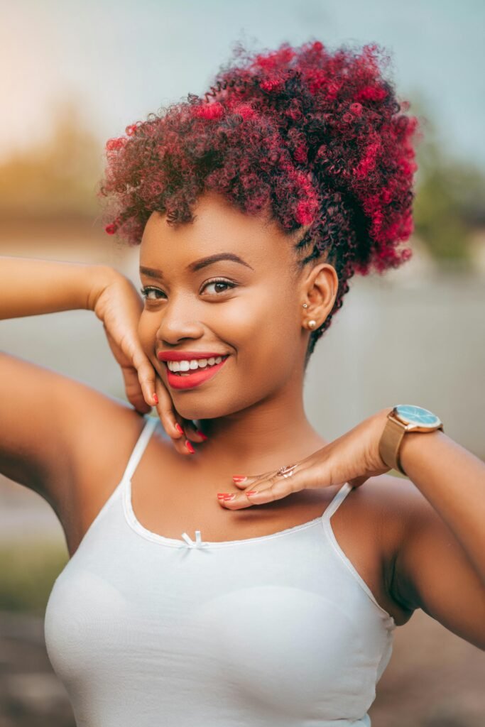 mulher preta linda sorrindo maquiada cabelos crespos presos com penteado estiloso mexas de vermelho blusinha branca relógio no braco