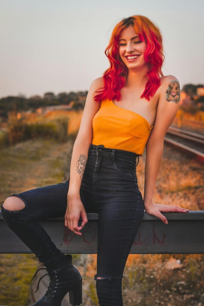 foto de mulher branca sorrindo cabelos longos lisos pintados de vermelho red calca jeans blusinha estilosa amarela tatuagens nos bracos
