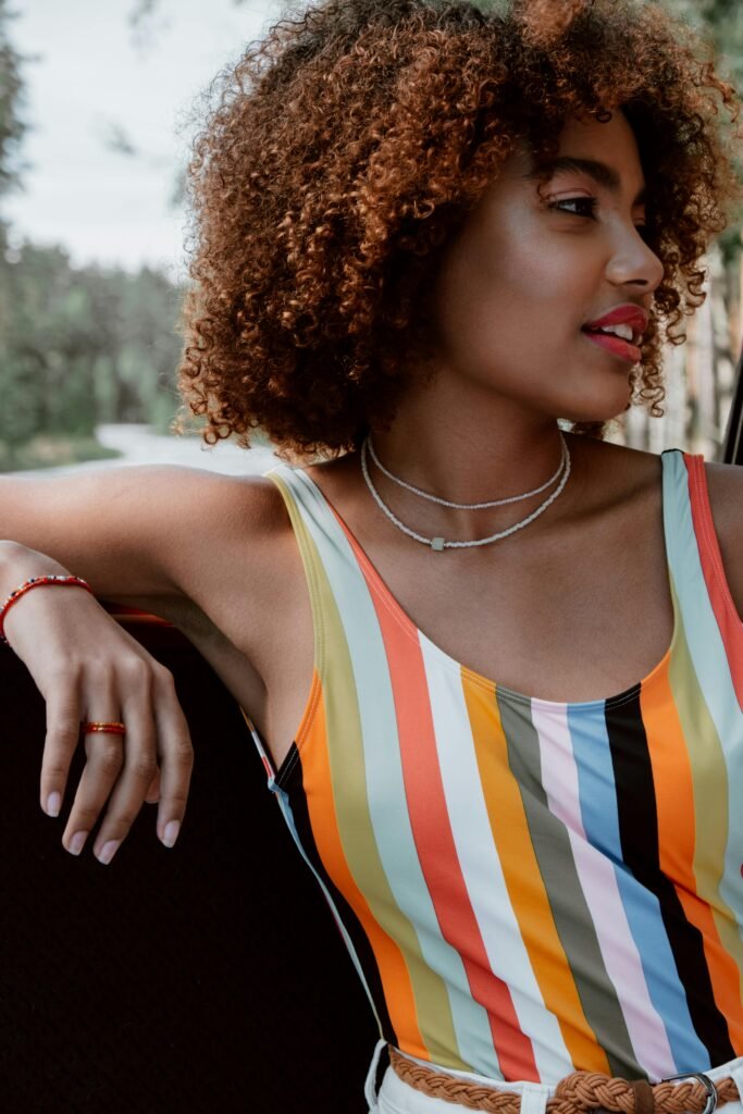 foto de mulher preta linda jovem leve sorriso cabelos cacheados 3c dois colar no pescoço blusinha listrada varias cores anel no dedo