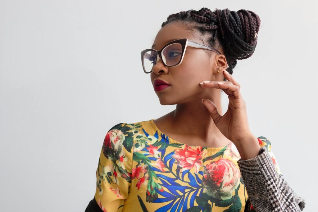 foto de mulher preta linda cabelos transados presos french hair pin usando óculos estiloso leve maquiagem uma das mãos no rosto