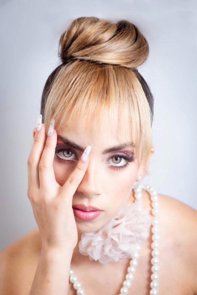 mulher loira linda olhos verdes claros maquiada uma das mãos no rosto cabelo preso penteado coque com franja