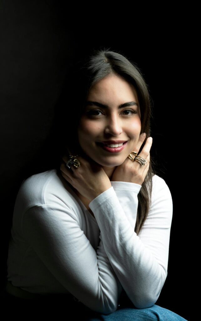 mulher morena de perfil sorrindo leve maquiagem cabelos lisos longos pretos anéis de ouro nos dedos blusinha branca