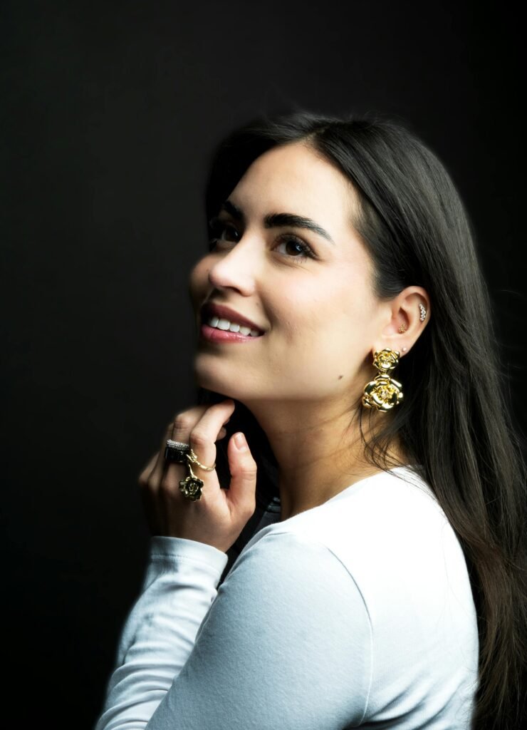 foto de perfil de mulher morena cabelos lisos pretos esta sorrindo brinco grande anéis e uma das mãos no queixo blusinha branca