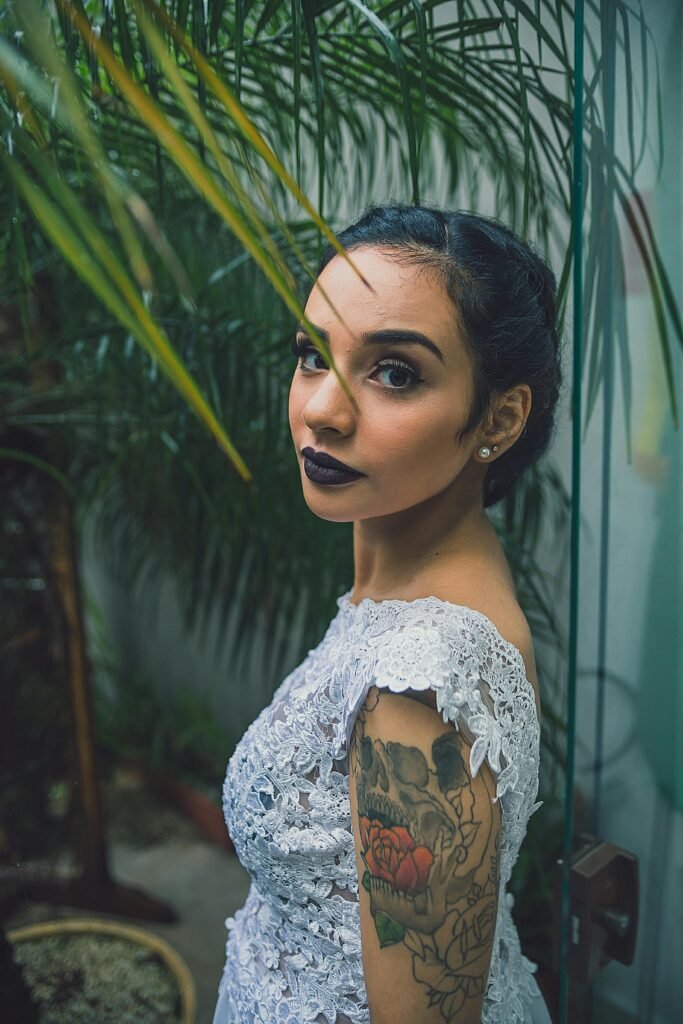 mulher branca cabelos lisos presos dois brincos pequenos perolas batom preto maquiada vestido estiloso branco tatuagem no braco