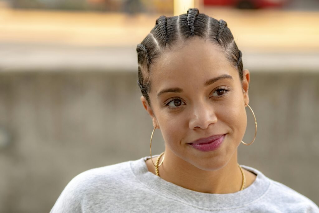 mulher com leve sorriso leve maquiagem brincos argolas grandes penteados no cabelo com varias trancas paralelas presas blusa cinza