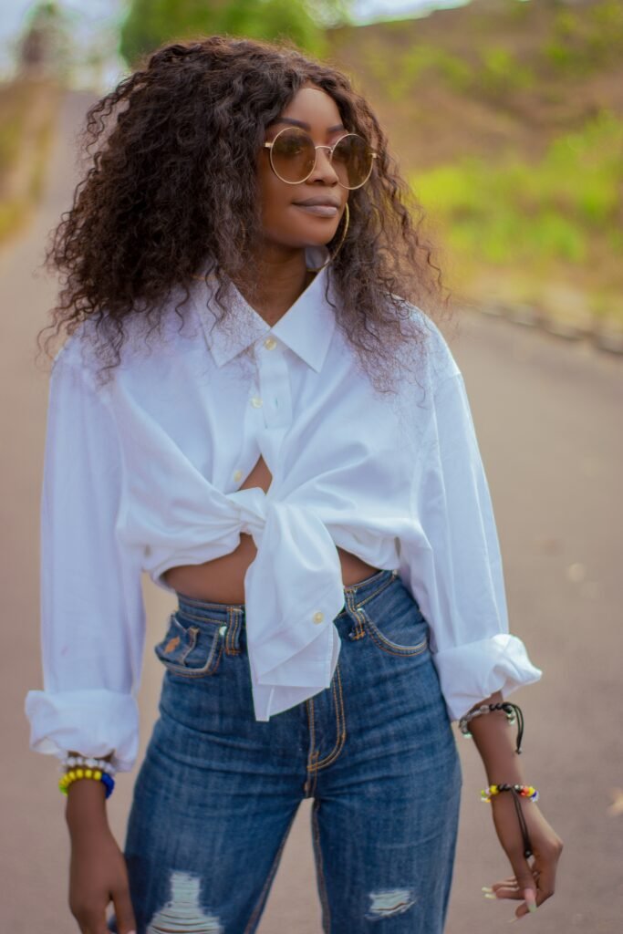 mulher com cabelos crespos grandes volumoso oculos redondo grande estiloso brincos argolas grandes camisa social branca com laco estiloso calca jeans azul rasgada pulseiras coloridas nos bracos