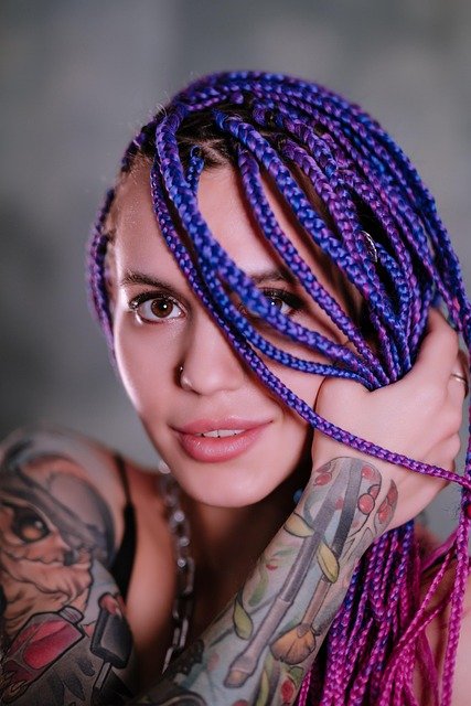 foto de perfil de mulher sorrindo com cabelos com varias trancas francesas na cor azul violeta uma das mãos segurando os cabelos tatuagens nos bracos