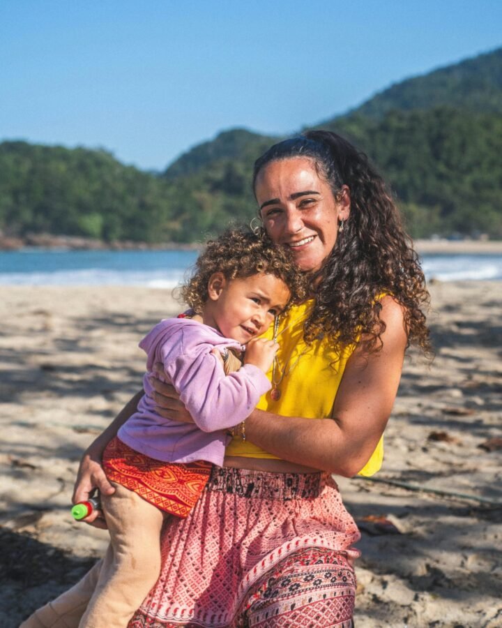 mae sorrindo feliz segurando sua filha pequena no colo também sorrindo estão na praia foto de Nando Martins unsplash.com