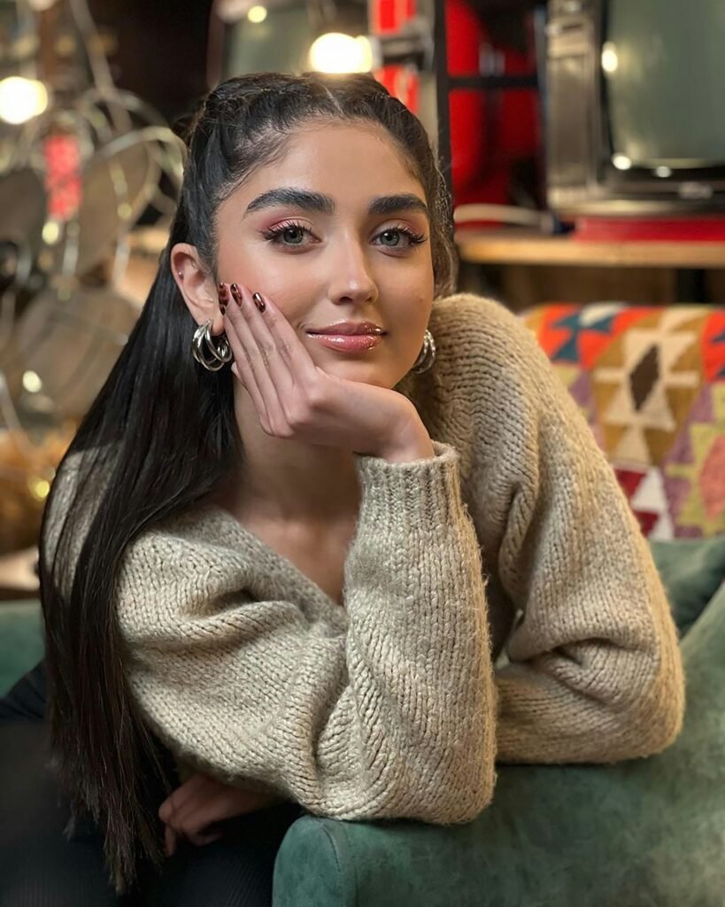 mulher maquiada leve sorriso cabelos longos lisos pretos brincos argolas prateados blusa bege uma das mãos no rosto